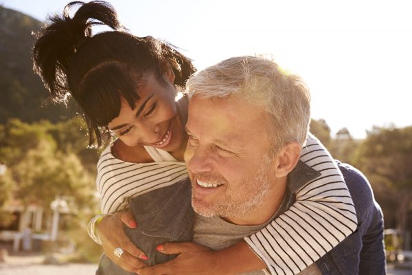 Man giving his girlfriend a piggyback ride outside, while both are laughing and smiling at each other